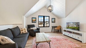 Un habitación tipo loft con techo inclinado, un ventanal con vistas, un sofá, una silla, una mesa y un televisor sobre un pedestal.