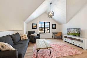 Ein Loft-Zimmer mit schrägem Dach, einem großen Fenster mit Aussicht, einer Couch, einem Sessel, einem Tisch und einem Fernseher auf einem Standfuß.