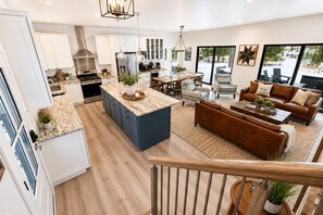 Een moderne keuken met een centraal eiland, een eetgedeelte met stoelen, een bank en uitzicht op de omgeving door grote ramen.