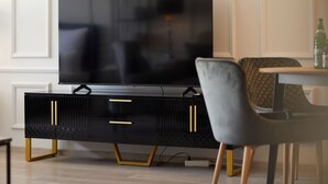 Ett modernt vardagsrum med en stor platt-TV placerad på en svart möbel för underhållningsutrustning med guldfärgade detaljer, en grå klädd fåtölj och ett litet sidobord.