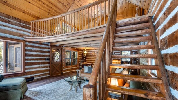 Interno di una baita in legno con scala centrale, camino e zona salotto accogliente.