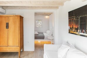 A modern interior with a wooden wardrobe, a white sofa, a bed with white bedding, and a painting of a classical scene. - J’adore Mykonos Villa – Iconic Pool & Views (MYKONOS)
