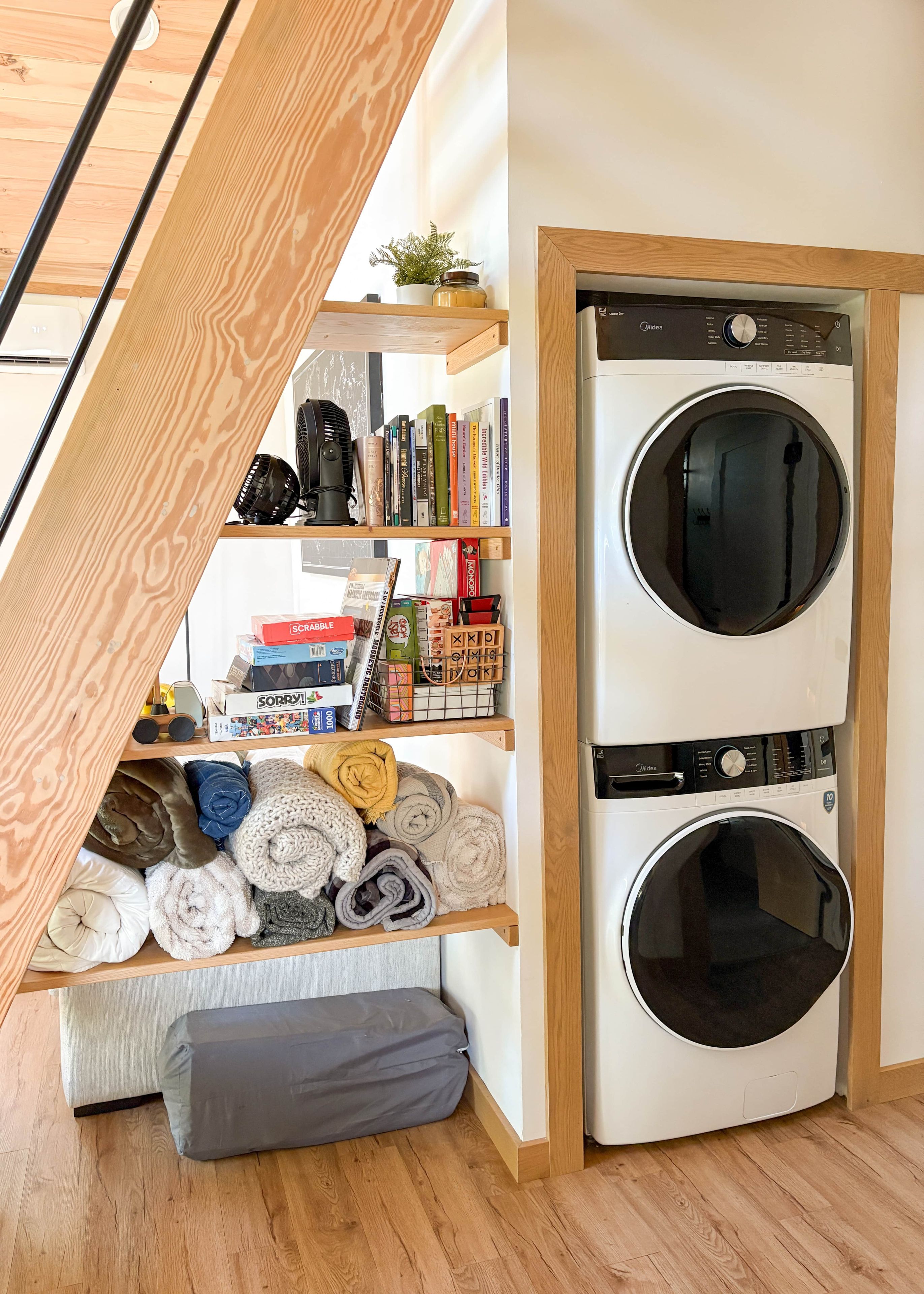 Ein Waschraum mit einer Frontlader-Waschmaschine und einem Trockner, ein Holzregal mit Büchern und gefalteten Handtüchern sowie eine kleine Pflanze auf dem Regal.