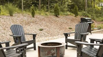 Una chimenea circular rodeada de sillas Adirondack en un patio con un sendero de grava y árboles al fondo.