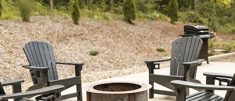 Ein runder Feuerkorb, umgeben von Adirondack-Stühlen auf einer Terrasse mit Kiesweg und Bäumen im Hintergrund.