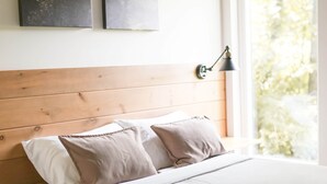 Quarto com cabeceira de madeira, cama com roupa de cama branca, duas almofadas, abajur e uma obra de arte na parede.