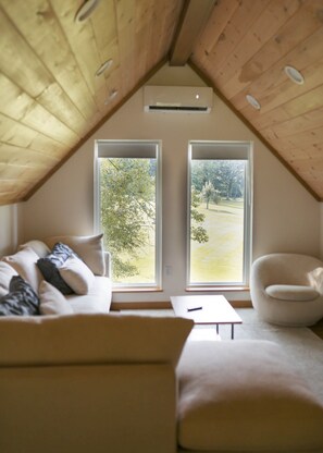 A cozy attic room with a sloped ceiling, a sofa, a chair, and two large windows offering a view of a grassy area.