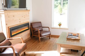 Una habitación con chimenea, un televisor montado sobre ella, un sillón de cuero marrón, una mesa de madera con una bandeja y una ventana con vistas a la vegetación.