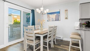 A dining area with a table and chairs, a chandelier, a painting, and a view through a sliding door to the outside. - Treasure’s Cove at Seaway | Steps to the Beach (Ocean City)