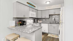 A modern kitchen with white cabinets, a granite countertop, a white stove, and a microwave. There are two stools and a colorful rug on the floor. - Treasure’s Cove at Seaway | Steps to the Beach (Ocean City)