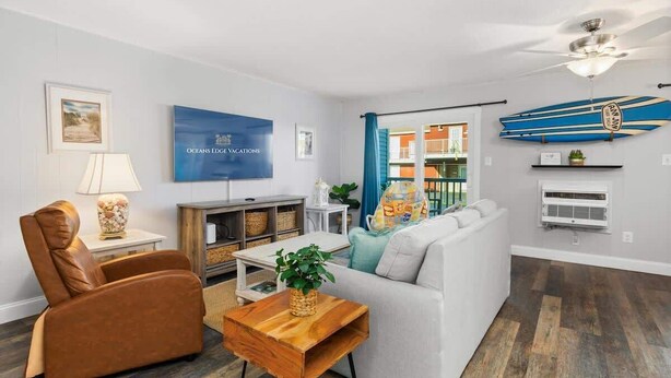 A modern living room with a sofa, armchair, coffee table, and a television. There is a ceiling fan, a wall-mounted surfboard decoration, and a framed picture on the wall. - Treasure’s Cove at Seaway | Steps to the Beach (Ocean City)