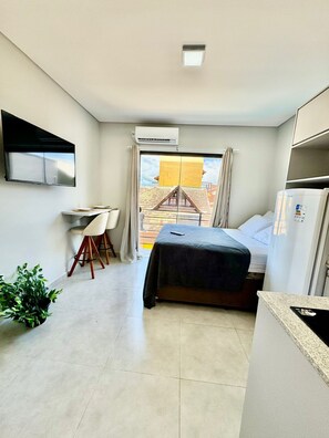 A compact hotel room with a bed, a desk, and a chair. There is a window with curtains, an air conditioning unit, and a television mounted on the wall. - Estudio Campeche (Florianópolis)