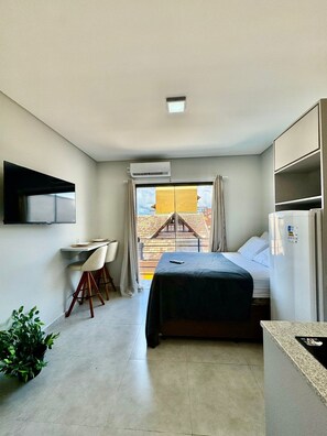 A compact hotel room with a bed, a desk with chairs, a TV mounted on the wall, and a window with curtains. - Estudio Campeche (Florianópolis)