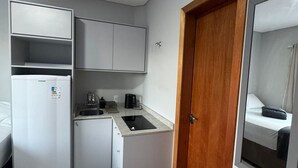 Une cuisine compacte équipée d’un réfrigérateur, d’un évier et d’un plan de travail, et une porte en bois donnant sur une chambre.