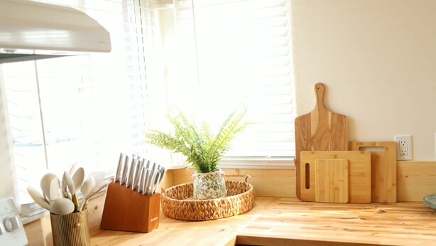 Una cocina con mostrador de madera, una planta en una cesta tejida, un bloque de cuchillos y una tabla de cortar.