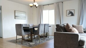 Una sala de estar con una mesa de comedor, sillas grises, un sofá gris, una araña y dos cuadros en las paredes.