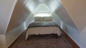Un dormitorio bajo y estrecho en un ático con una cama, una ventana y una alfombra estampada.