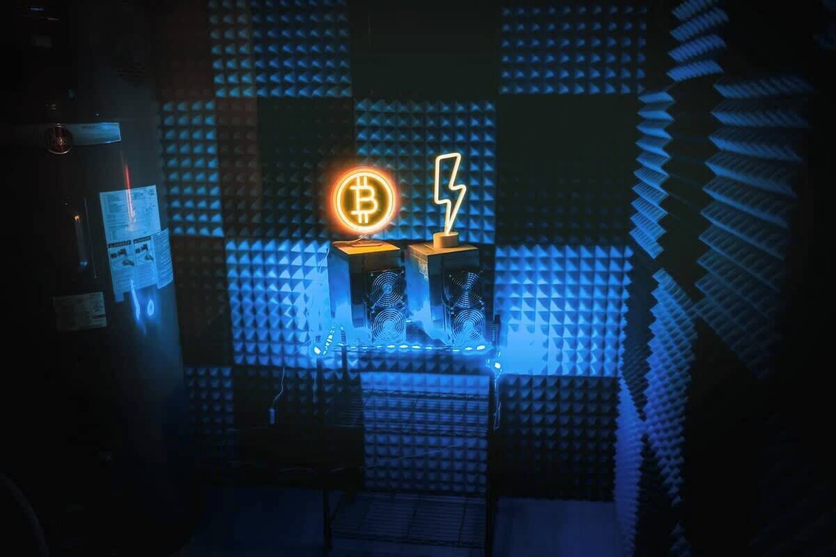 A room with a blue acoustic panel wall, a neon sign with a Bitcoin symbol and lightning bolt, and a small table with a chair.
