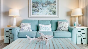 A teal-colored sofa with three cushions, one labeled "Ocean," flanked by two nightstands with lamps and decorative items. - Wake Up to Waves | Stunning Oceanfront Condo (North Myrtle Beach)