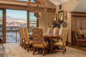 A dining room with a large wooden table and upholstered chairs, a chandelier, and a view of a snowy landscape through large windows. - Four Bedroom Penthouse in Teton Village (Teton Village)