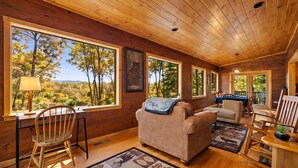 Interior acogedor de una cabaña con paredes de madera, ventanas amplias con vistas a árboles y montañas, un sofá, una mesa de comedor y una silla.
