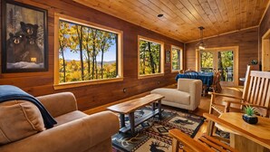 Interior acogedor de una cabaña con paredes de madera, ventanas amplias que ofrecen vistas a árboles otoñales, una zona de comedor con mesa y sillas, y una chimenea.