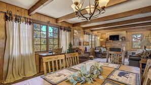 A rustic cabin interior with a wooden dining table, chairs, a chandelier, and a fireplace.