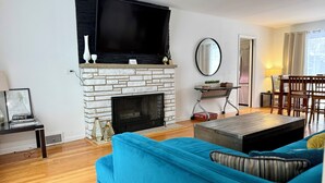 A living room with a fireplace, a flat-screen TV mounted above it, a blue sofa, and a dining area with wooden chairs and a table. - Cozy Estate in charming Willow Springs 20 minutes from Downtown and Beaches (Willow Springs)