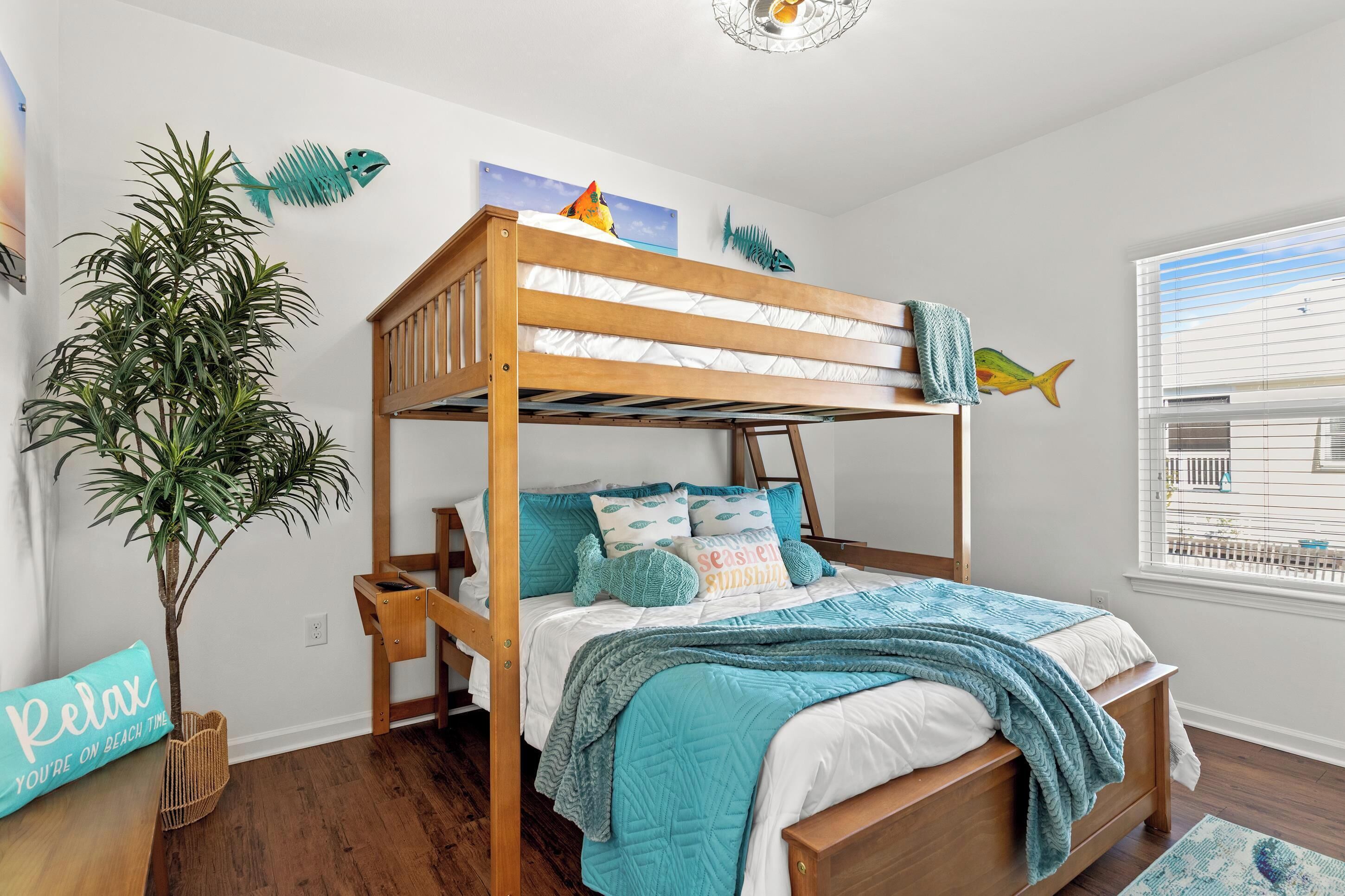 Una cama tipo litera con estructura de madera, ropa de cama blanca y detalles color turquesa. Una planta en maceta está al lado de la cama, y se ve una ventana con persianas.