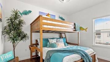 Una cama tipo litera con estructura de madera, ropa de cama blanca y detalles color turquesa. Una planta en maceta está al lado de la cama, y se ve una ventana con persianas.