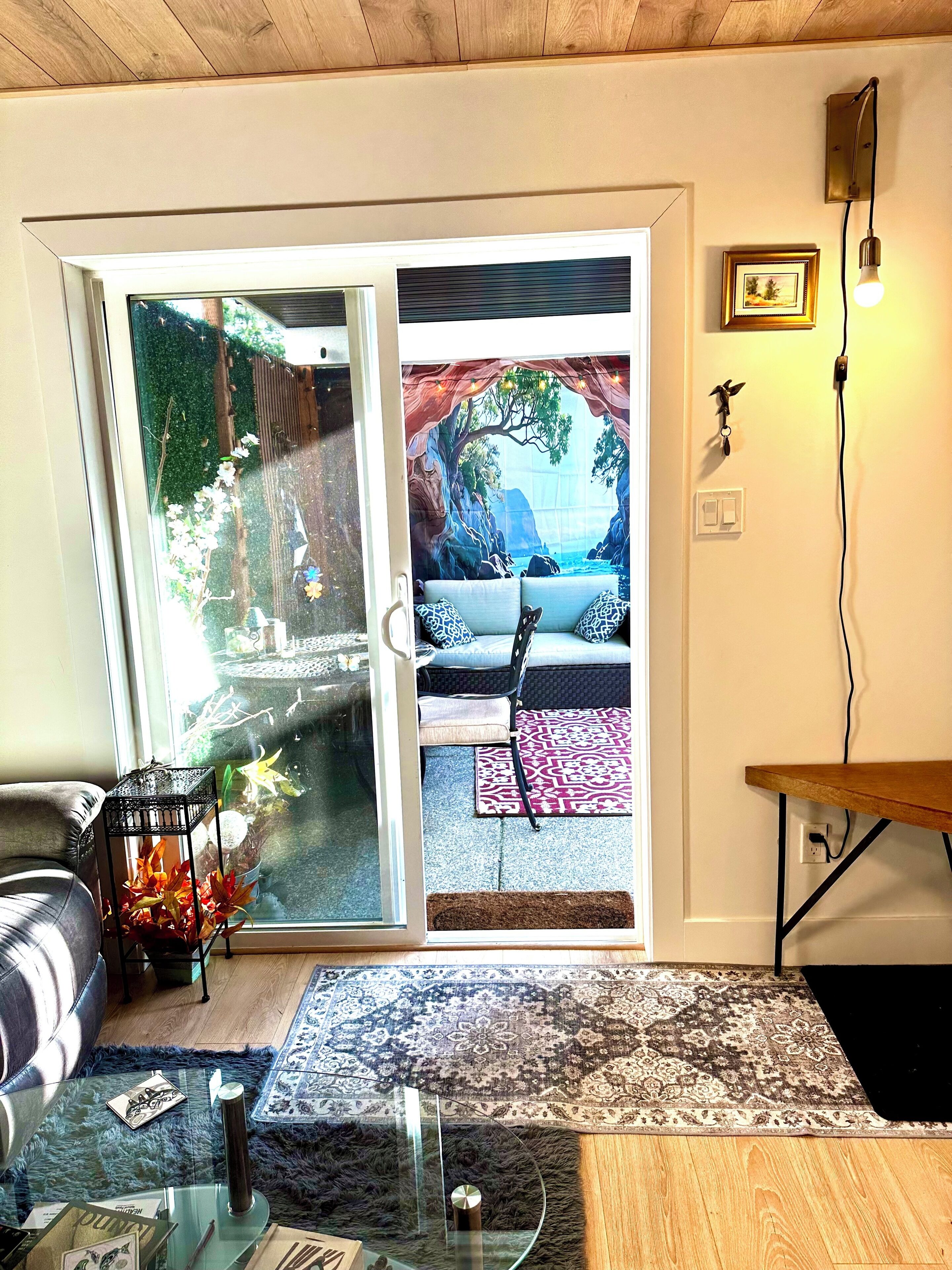 A living room with a glass coffee table, a sofa, a patterned rug, and a view through a sliding glass door to an outdoor area with a bench and a tree.