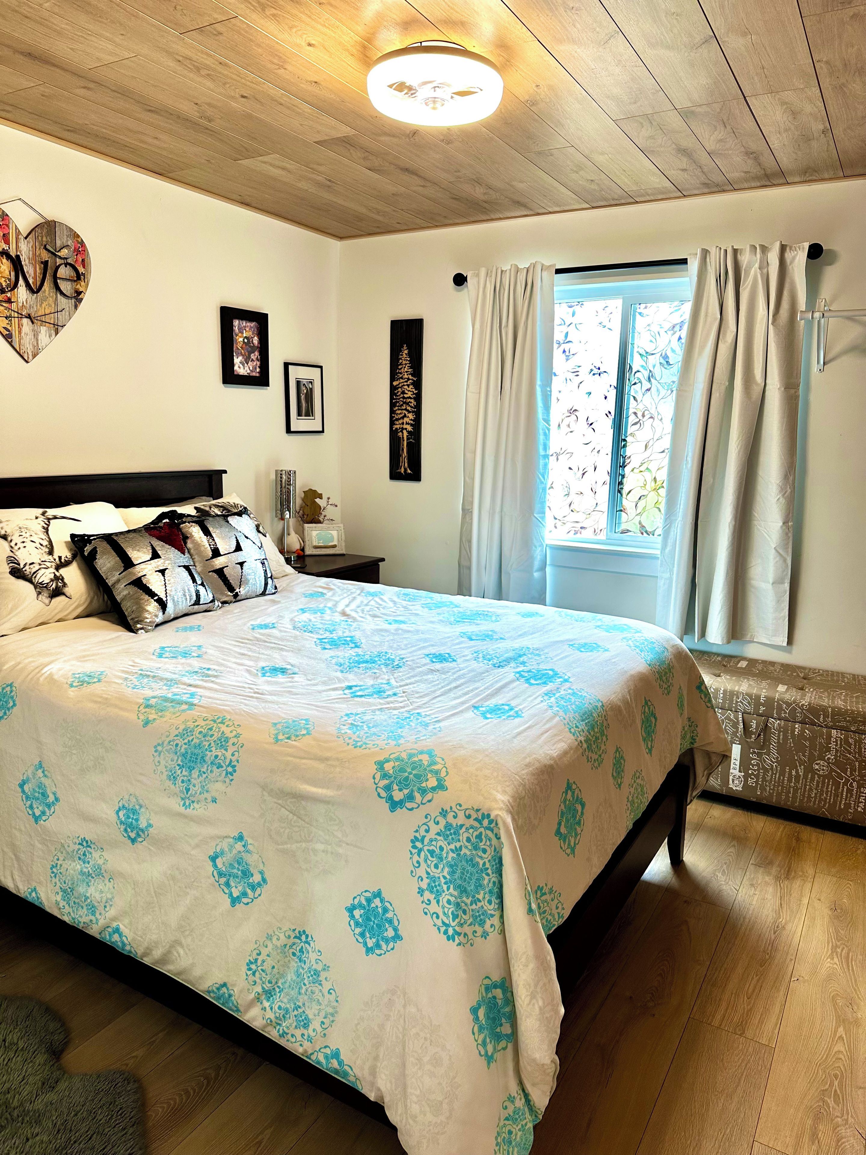 A bedroom with a bed, a window with curtains, a wall with a heart-shaped decoration, and a ceiling light.