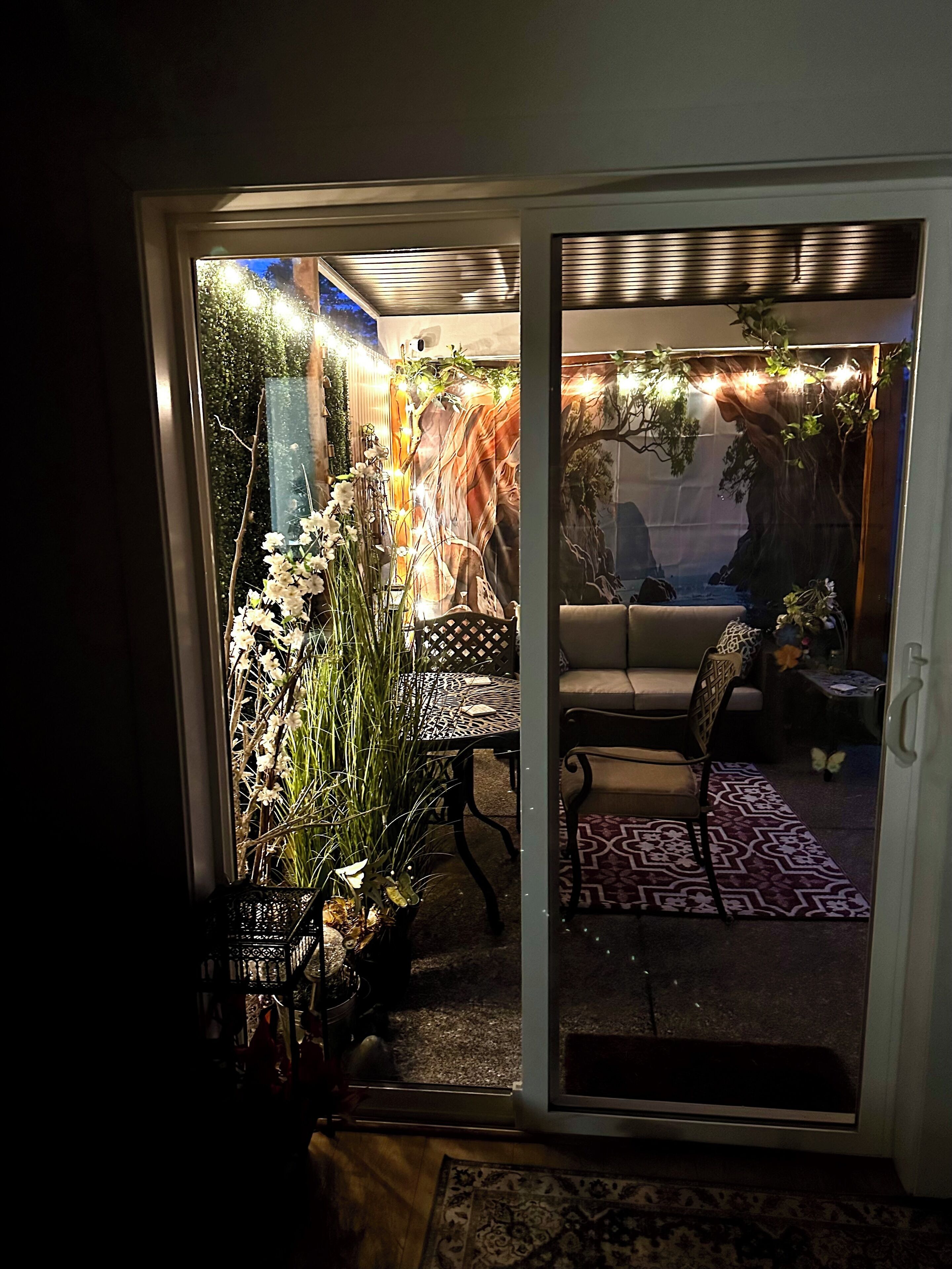 A cozy indoor patio with a sliding glass door leading to a garden area. There is a patterned rug, a cushioned seating area, and a table with a patterned tablecloth.