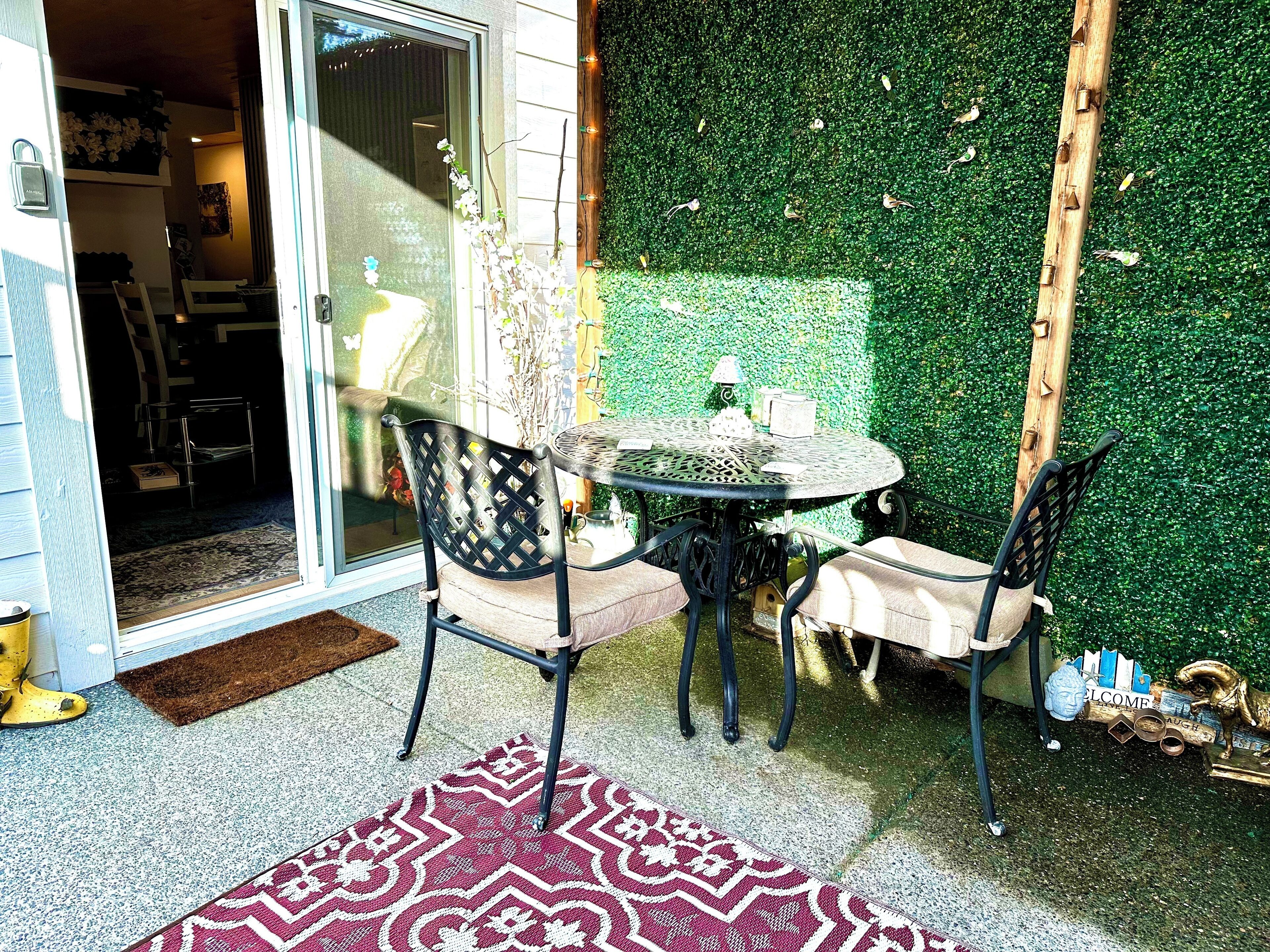A patio with a round table and two chairs, a patterned rug, and a green hedge wall.