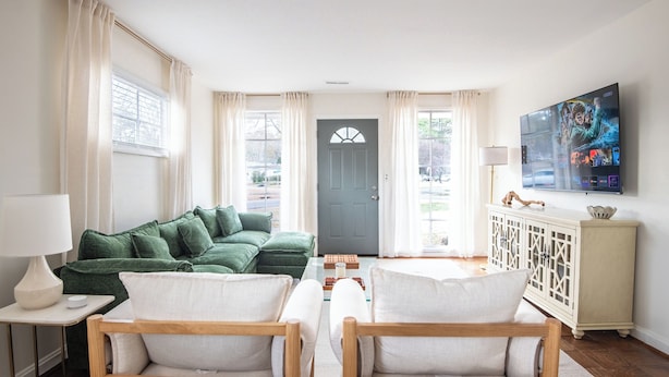 Un salon avec un canapé vert, deux fauteuils en bois, un meuble blanc et un téléviseur fixé au mur.
