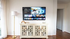 Un salon avec un téléviseur à écran plat fixé au mur, un meuble blanc avec des portes en verre, un lampadaire et un plancher en bois.