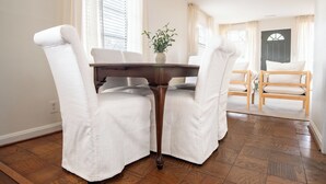 Un coin repas avec une table en bois et des chaises à revêtement blanc, une plante en pot posée sur la table, et, en arrière-plan, une porte et une fenêtre.