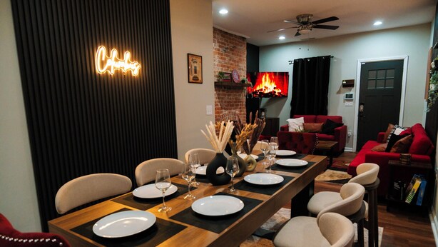 Un área de comedor con una larga mesa de madera dispuesta para seis personas, rodeada de sillas color beige. Se observa una chimenea con una repisa roja al fondo, y un letrero de neón en la pared que dice "Chic".