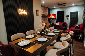 A dining area with a long wooden table set for six, surrounded by beige chairs. A fireplace with a red mantle is visible in the background, and a neon sign on the wall reads "Chic".