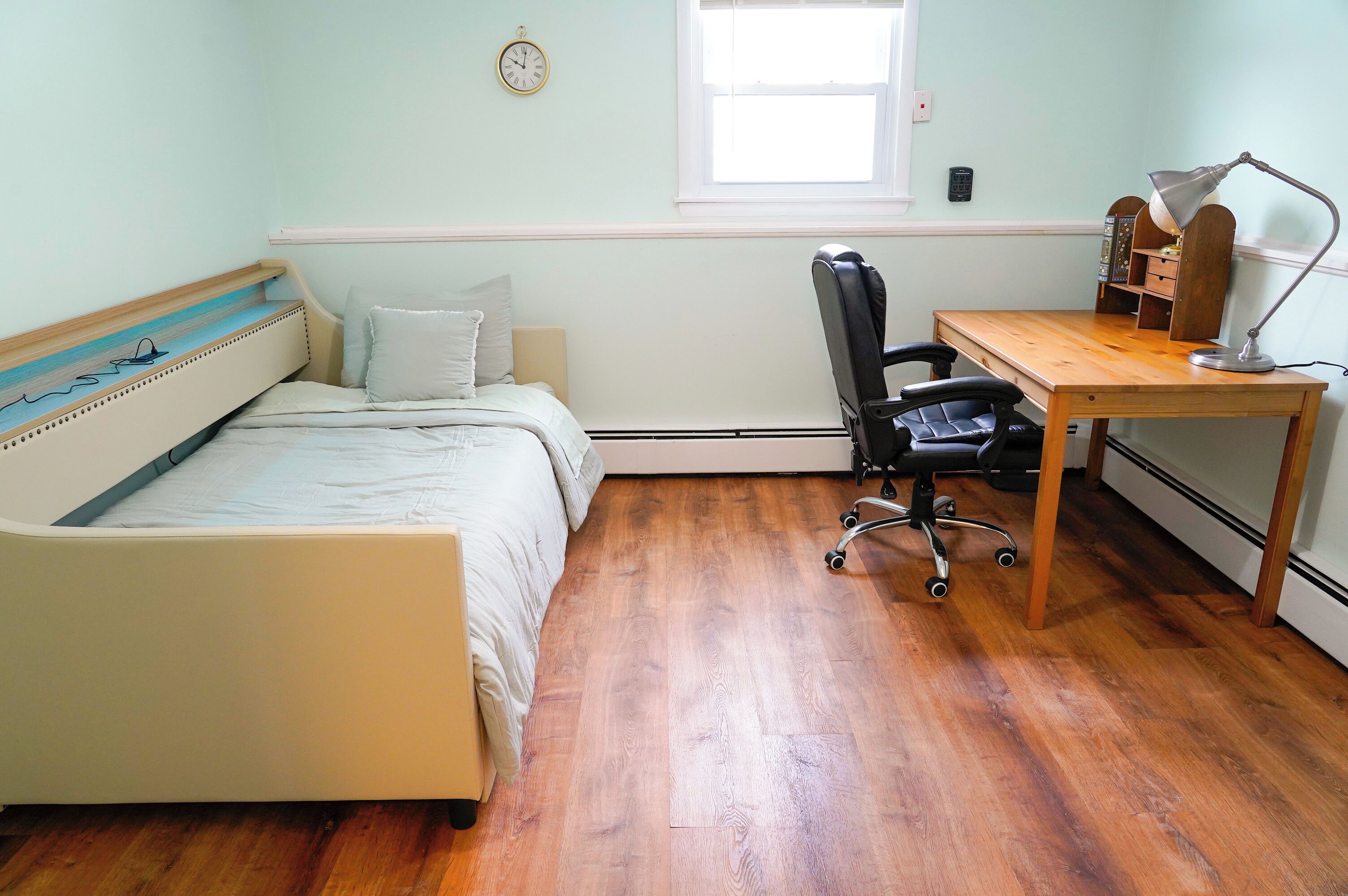 Een slaapkamer met een bed, een bureau, een stoel, een klok en een raam.