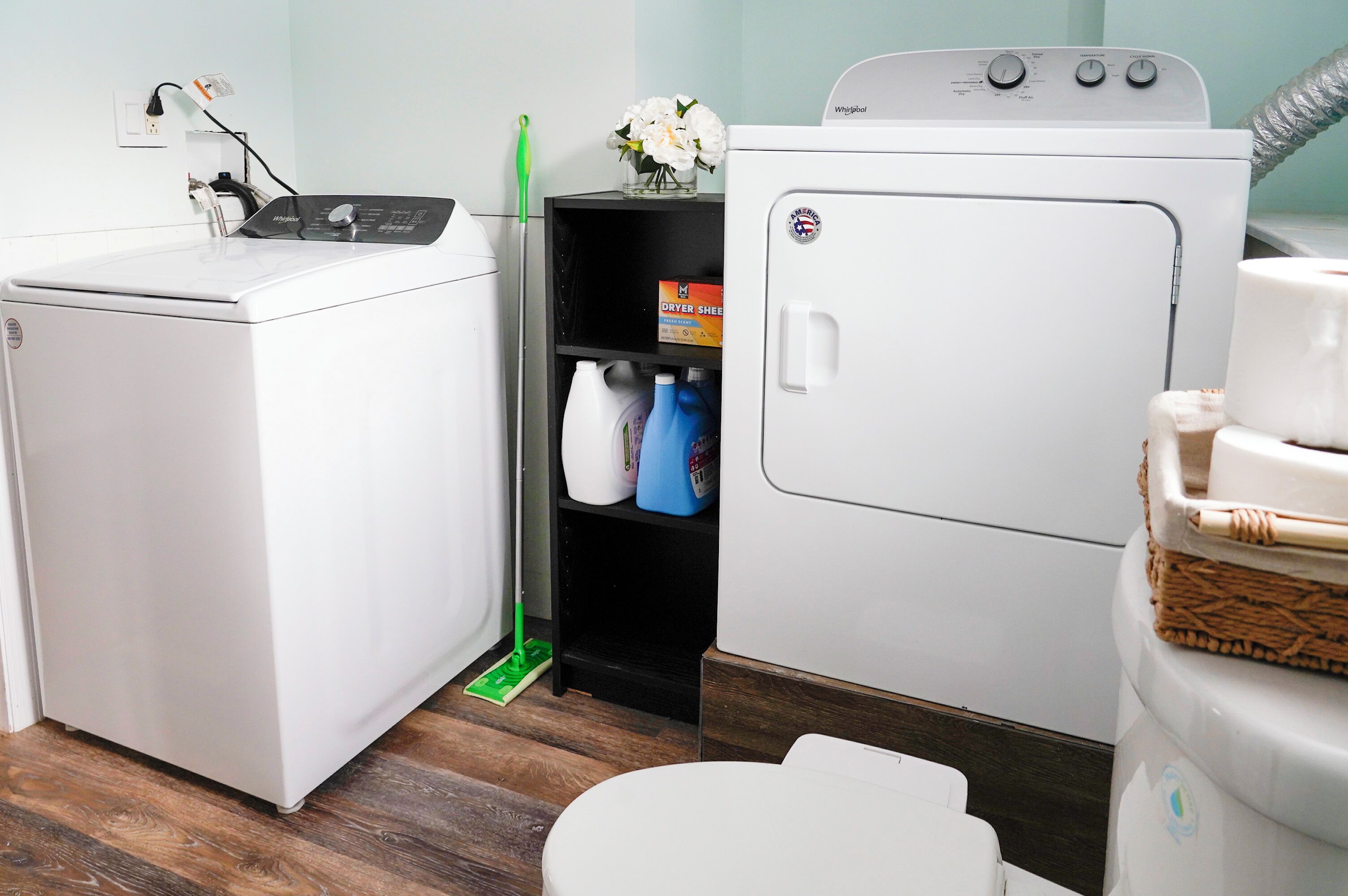 Une buanderie équipée d’un lave-linge et d’un sèche-linge à chargement frontal, d’une étagère contenant des produits de nettoyage et d’un panier en osier posé sur le sèche-linge.
