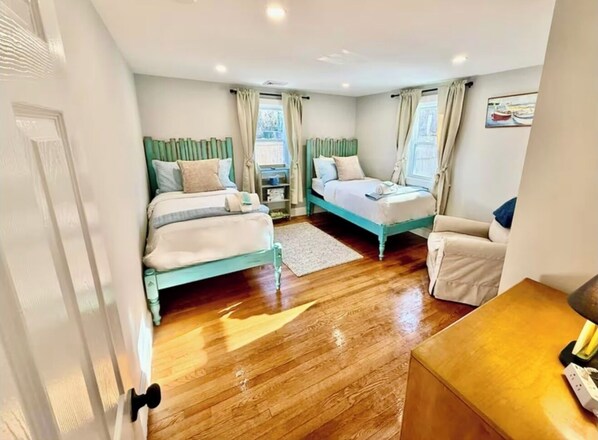 A bedroom with two beds, a wooden floor, a small table, and a window with curtains.