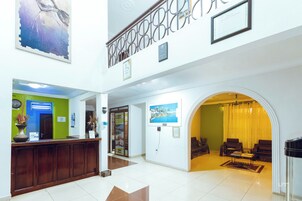 Un vestíbulo de hotel con mostrador de recepción, una pared verde, un cuadro grande y un área de estar con una cortina amarilla.