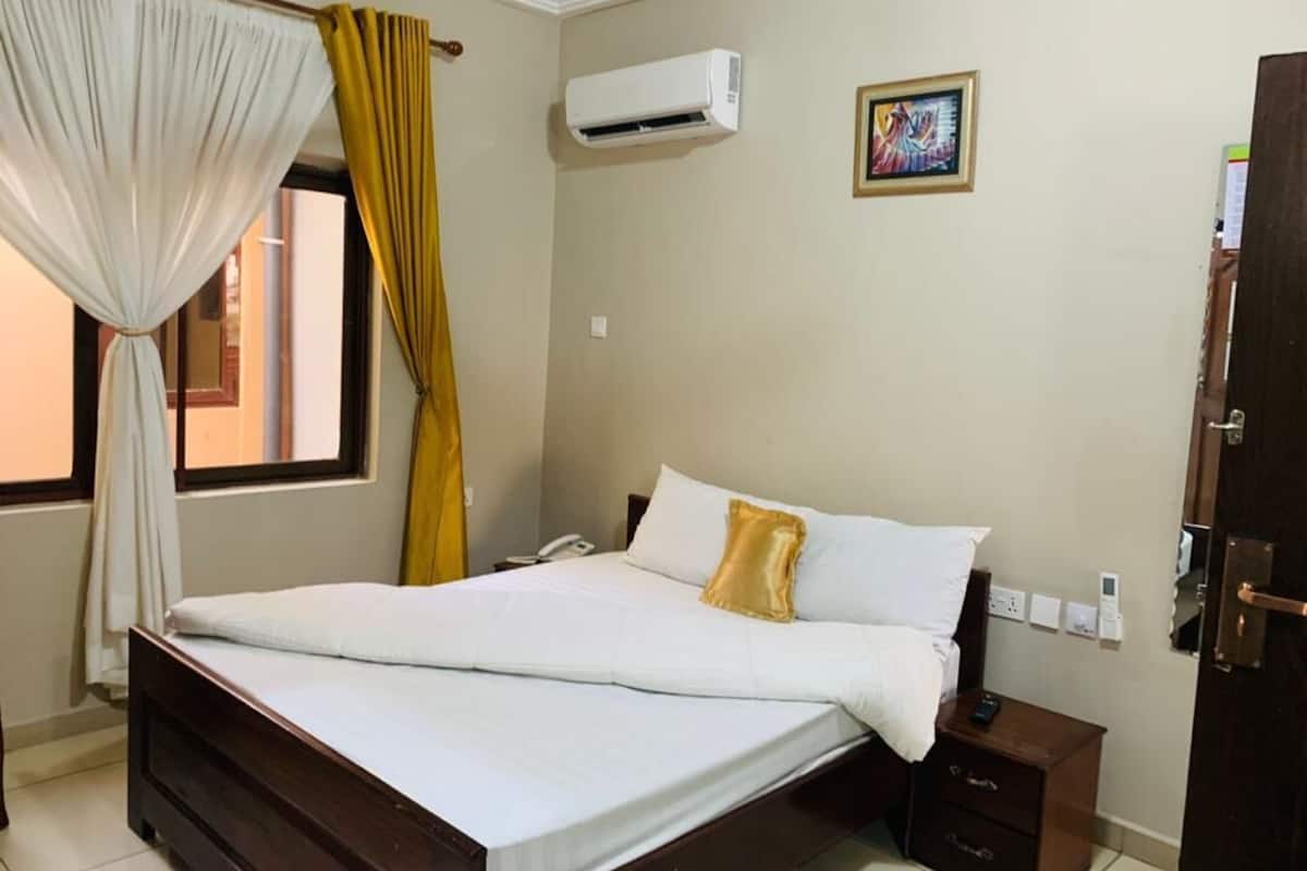 A bedroom with a bed, a bedside table, an air conditioning unit, a window with curtains, and a door.