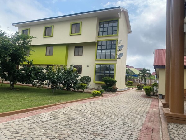 A two-story building with a green and white facade, satellite dishes, and a paved walkway leading to the entrance. - Excelsa Lodge (Ejisu)