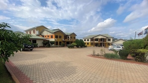 A large building with multiple sections, a paved driveway, and parked cars. - Excelsa Lodge (Ejisu)