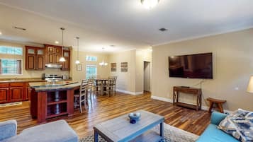 A modern kitchen with wooden cabinets, a dining area with a table and chairs, a living room with a sofa and a TV, and a hallway with a door.