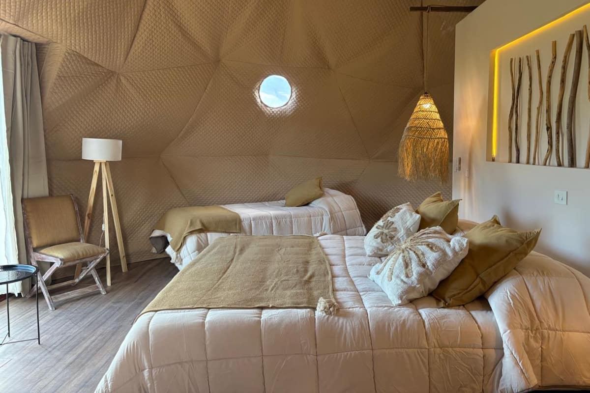 Une chambre à coucher avec un plafond au motif géométrique, une lucarne circulaire, un lit agrémenté de plusieurs coussins, une lampe sur pied et un luminaire fixé au mur.