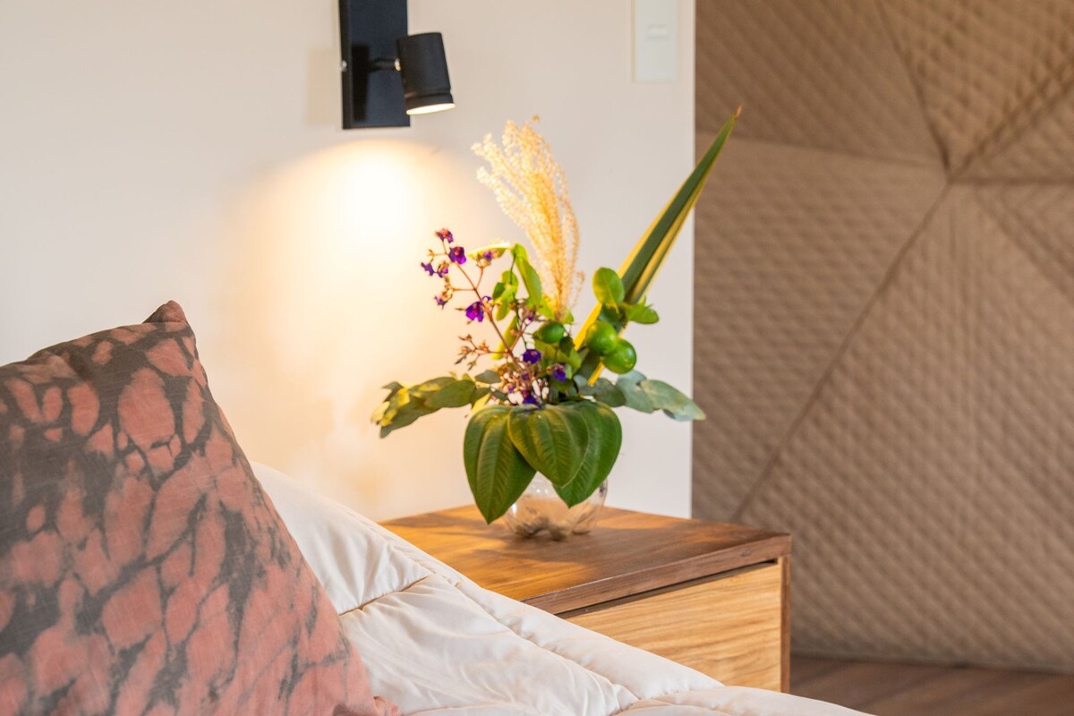 Un lit bien fait, agrémenté d’un coussin à motifs, une table de chevet avec un vase de fleurs et un luminaire fixé au mur.