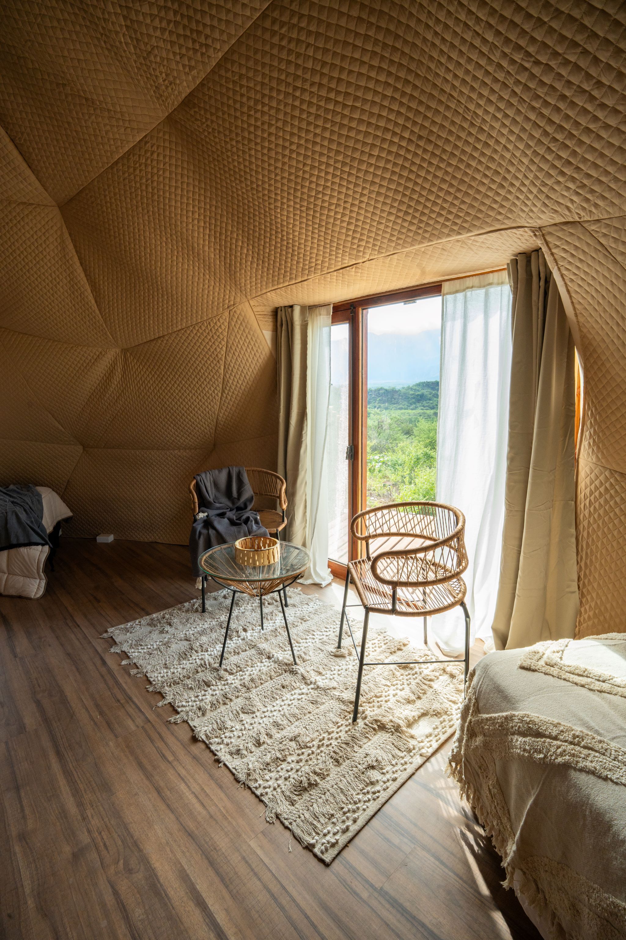A room with a geometric ceiling, a bed, a wicker chair, a glass table, and a view through a large window.